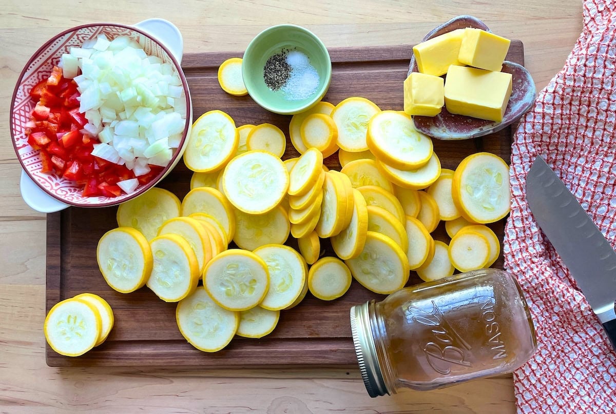 The ingredients in buttery squash on a cutting board - sliced yellow squash diced onion, red bell pepper, butter, salt, pepper, sugar, and chicken stock.