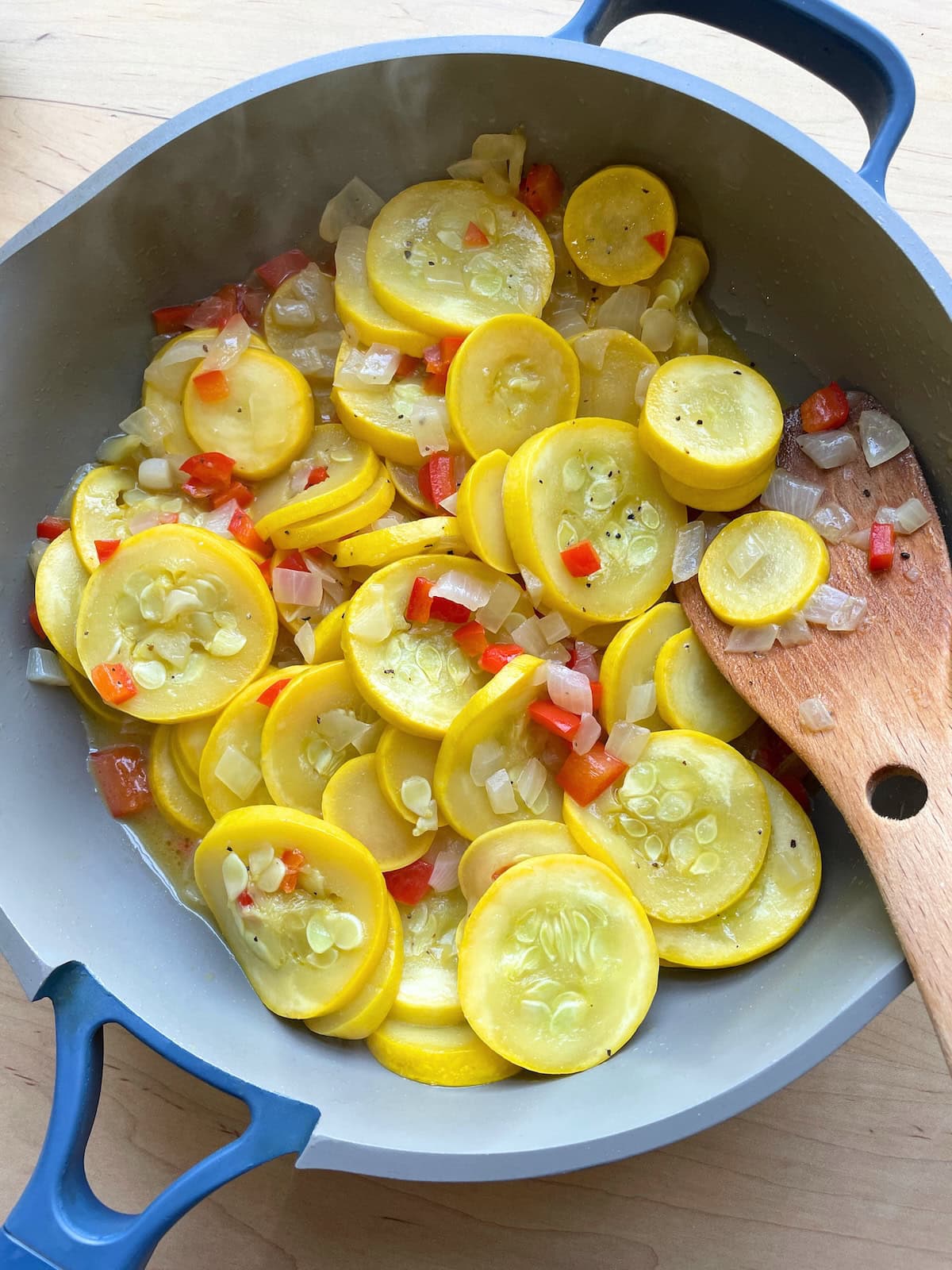 A blue and gray frying pan with cooked summer squash, red bell peppers, and onions in it with a wooden spoon.