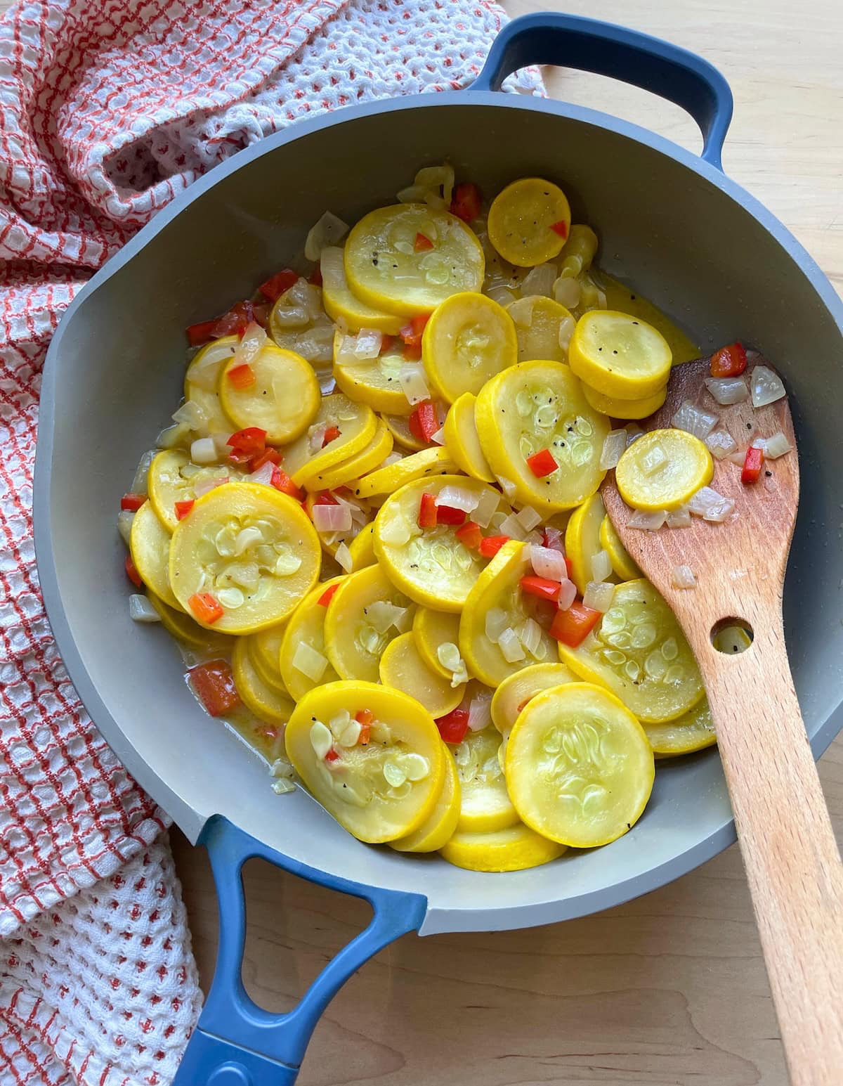 A blue and gray frying pan with buttery yellow summer squash and onions in it with a wooden spoon.