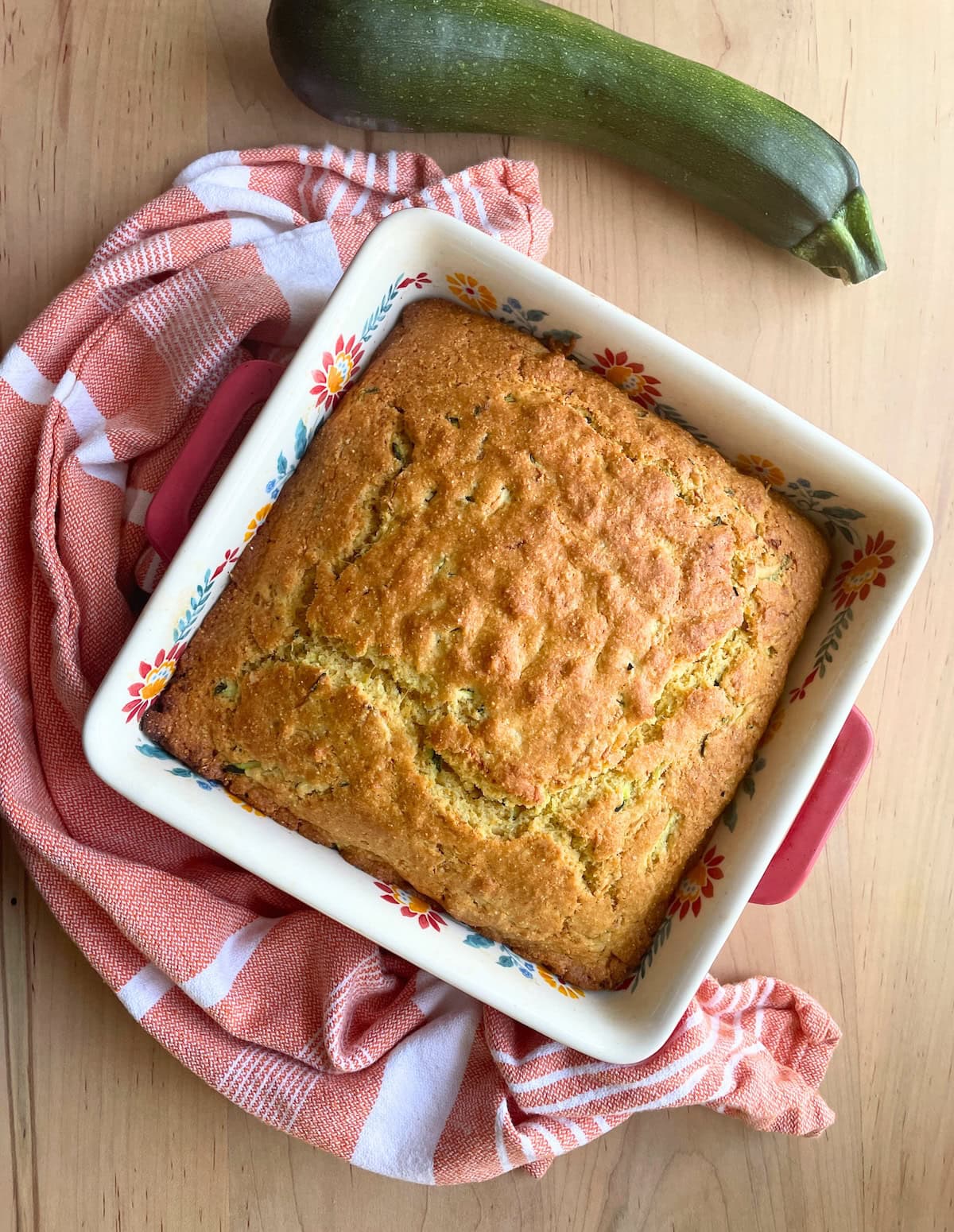 A square white baking dish with baked zucchini cornbread in it and a zucchini on the side.