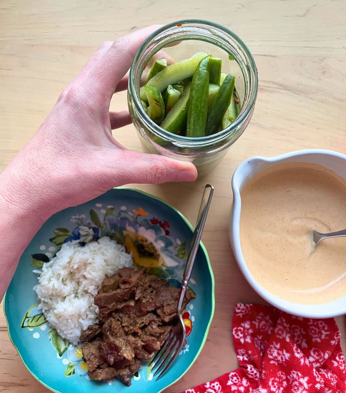 A bowl of rice with beef bulgogi, a bowl of Yum Yum sauce, and a hand holding a jar of Korean pickles.