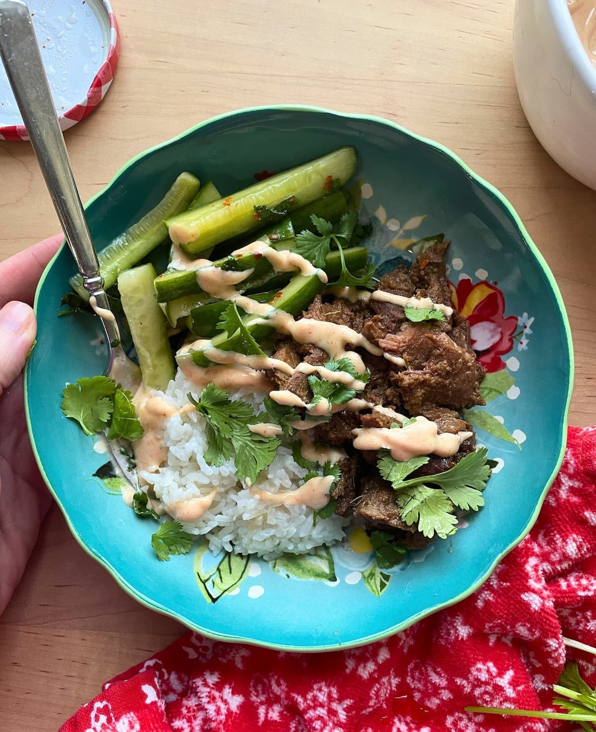 A hand holding a blue bowl with rice, bulgogi, Korean pickles, Yum Yum sauce, and cilantro.