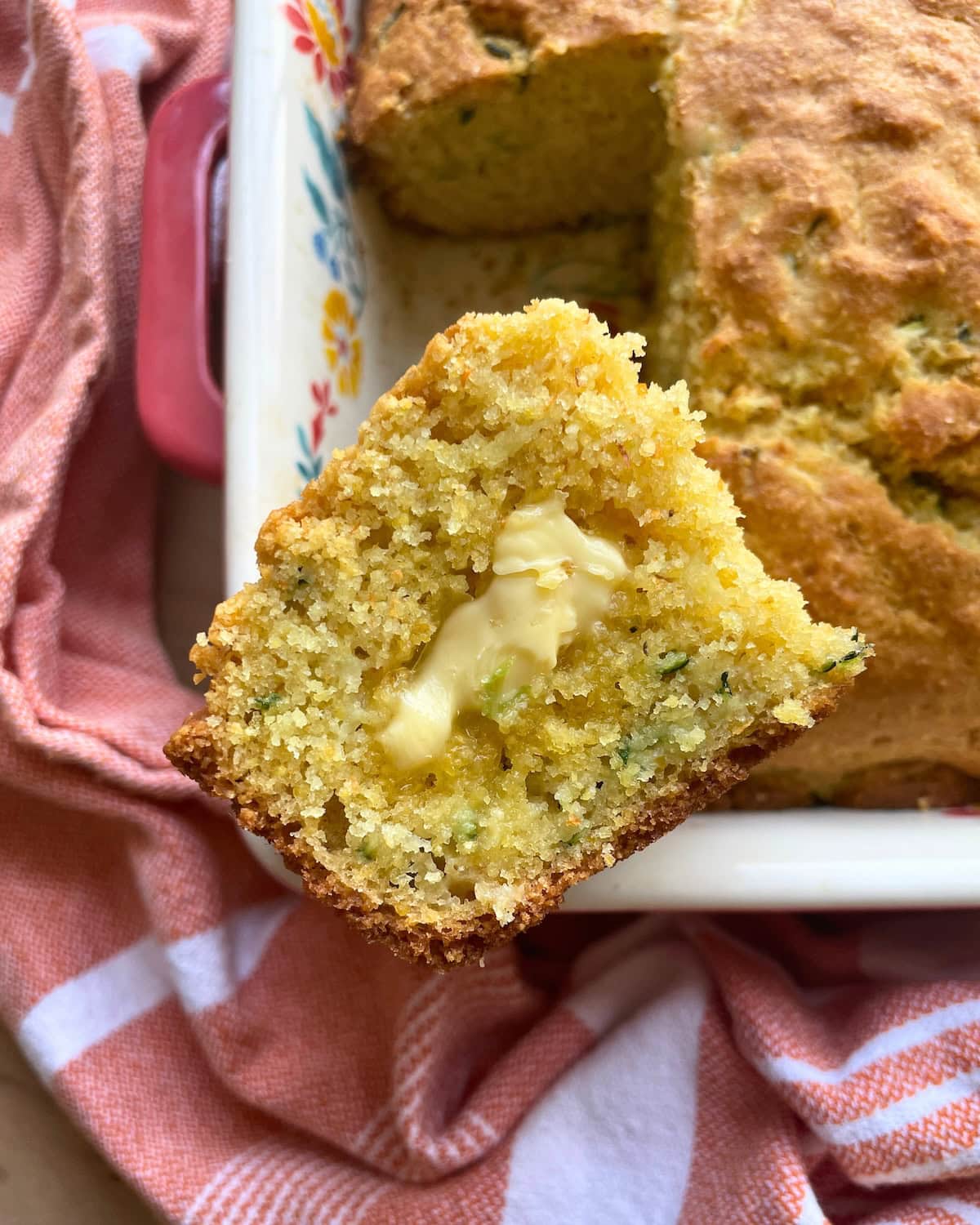 A slice of zucchini cornbread sitting on the edge of the pan of cornbread with honey butter melting on it.