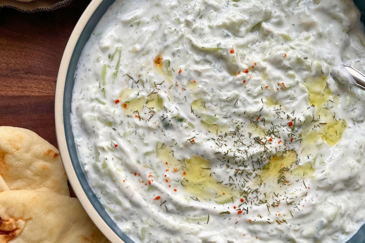 A close-up view of homemade tzatziki sauce garnished with dill and spices, served with slices of pita bread on the side.