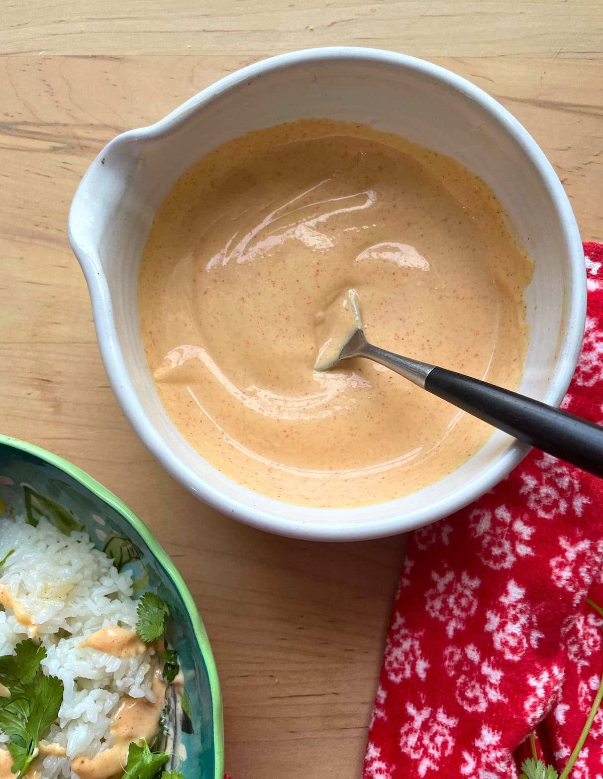 Yum yum sauce in a white bowl with a spoon next to a bowl of cilantro rice and a red and white towel.