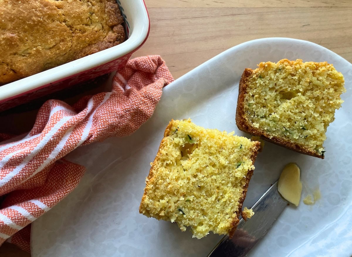 Two slices of zucchini cornbread on a white plate next to a buttered knife.