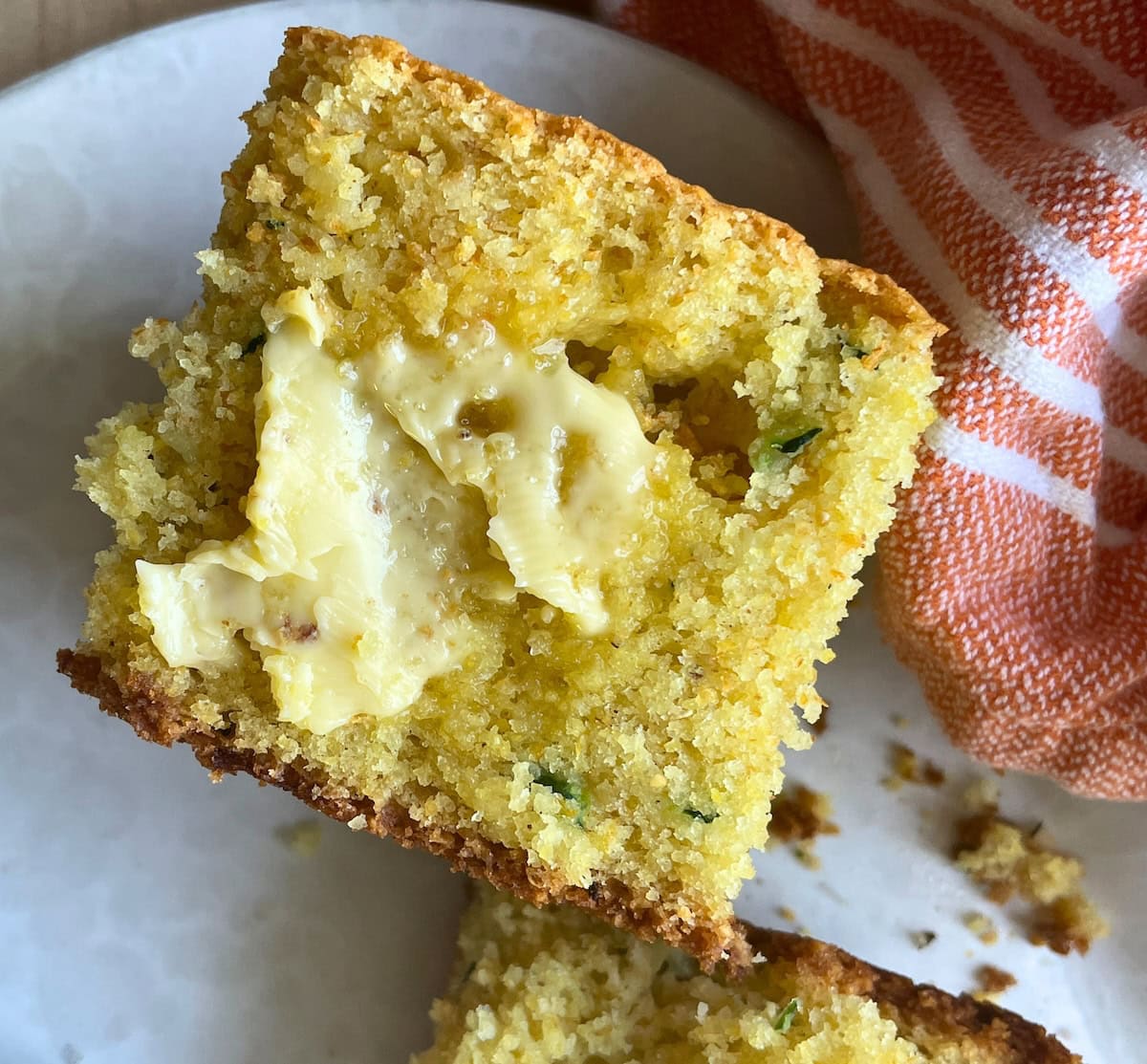 A slice of zucchini cornbread on a white plate with honey butter melting on it.