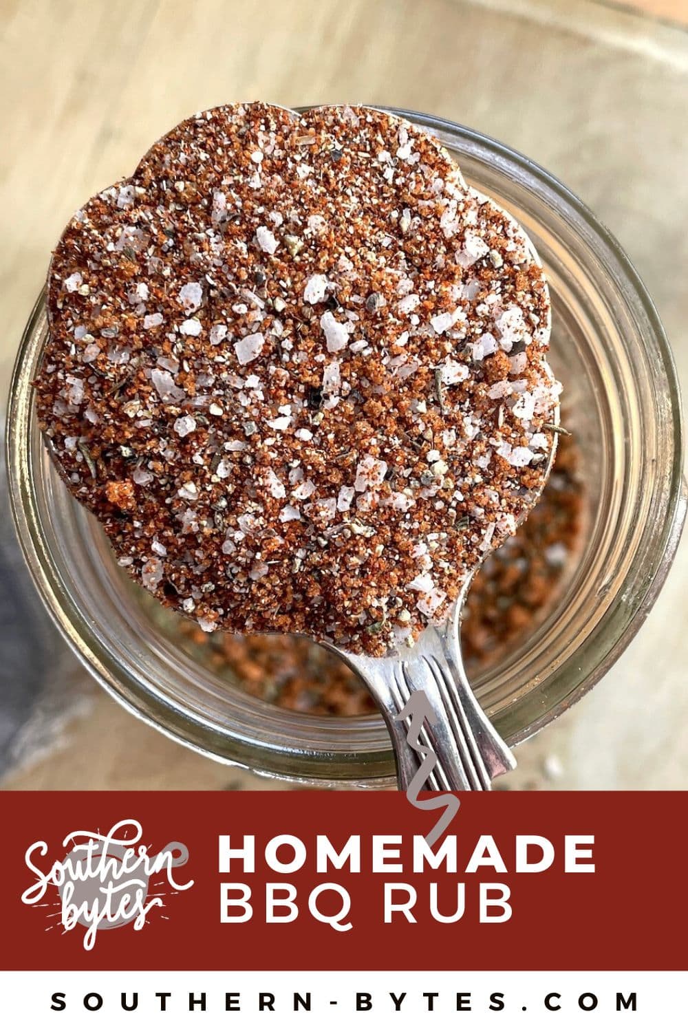 A pin image of a close-up view of a homemade BBQ spice rub in a glass jar with a spoonful of the mixture sitting on top.