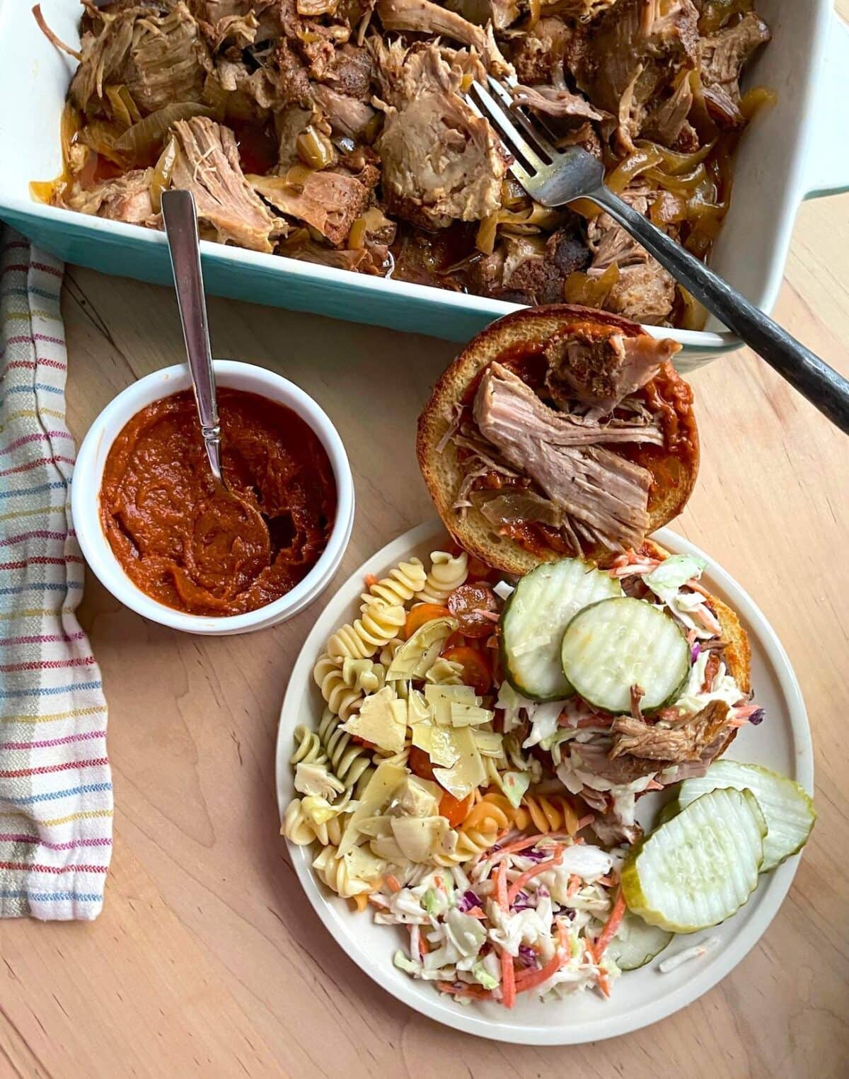 A plate of pasta salad, coleslaw, and a pulled pork sandwich with pickles, beside a bowl of BBQ sauce and a dish of shredded pork.