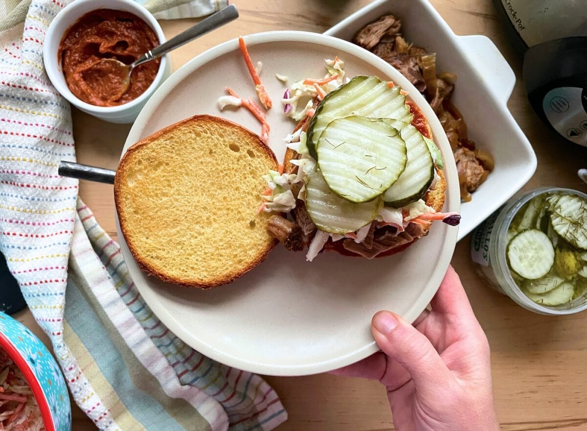 A hand holding a plate with a toasted bun, pulled pork, creamy coleslaw, and pickles on top, with condiments visible in the background.