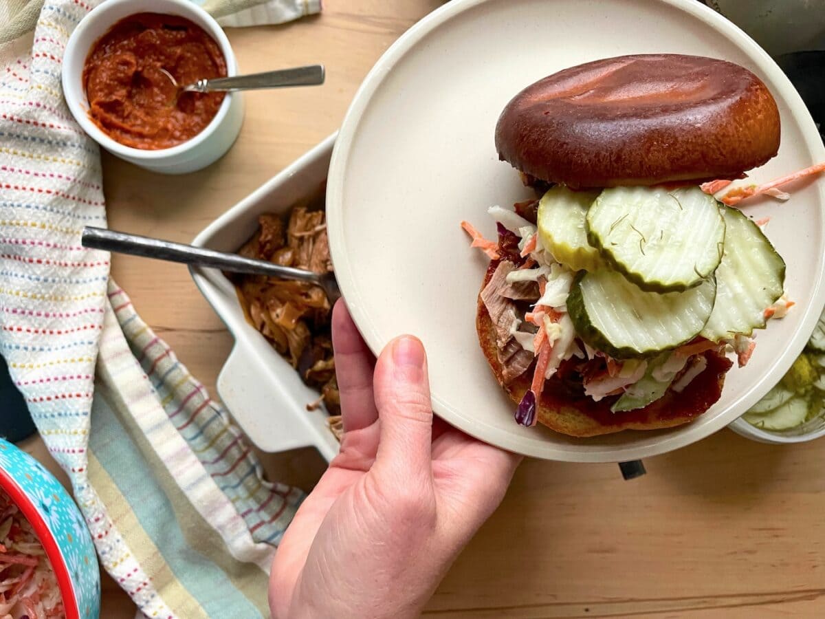 A person's hand holding a plate with a half-assembled pulled pork sandwich, pickles on top, next to bowls of additional ingredients.