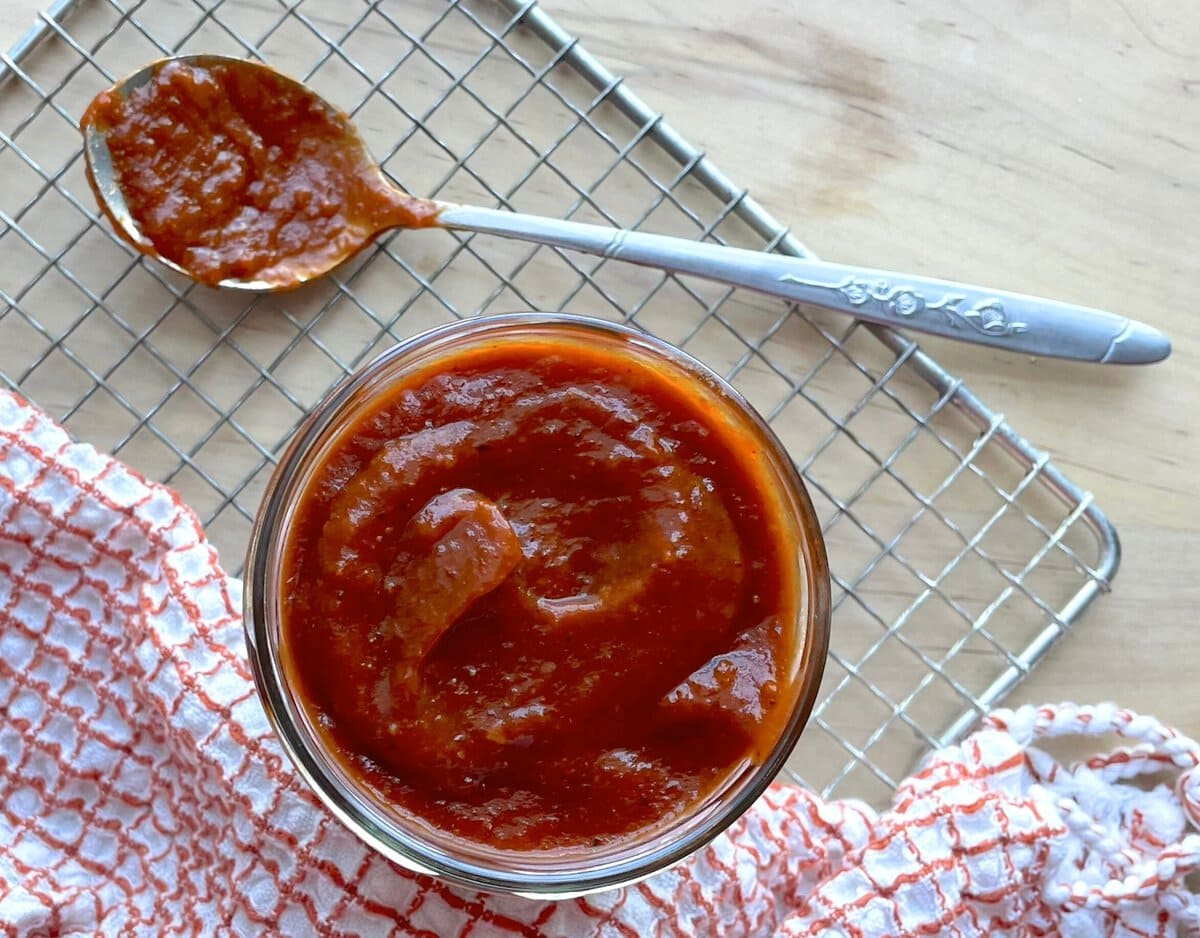 A jar of thick chipotle BBQ sauce next to a spoonful of sauce and a red and white dishcloth on a wooden surface.