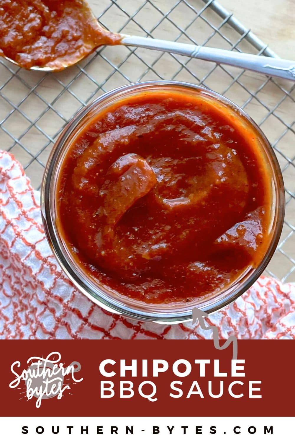 A pin image of an overhead view of a glass jar filled with chipotle BBQ sauce, a spoon with sauce on it, and a checkered cloth on the side.