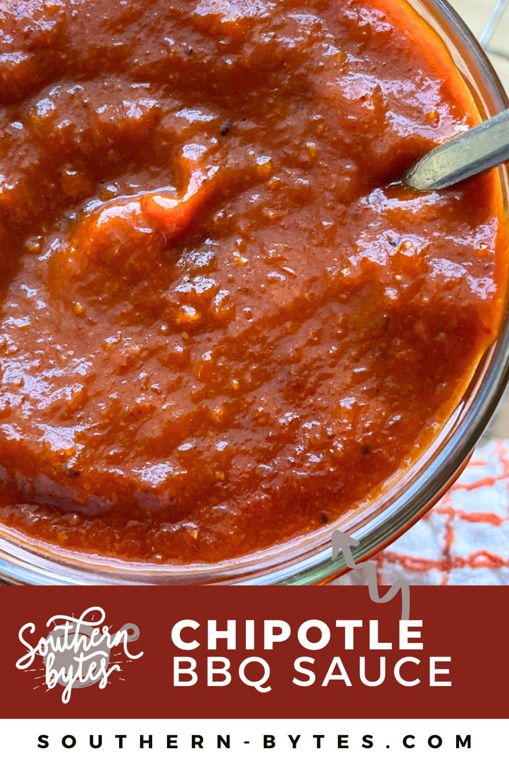 A pin image of a close-up view of homemade chipotle barbecue sauce in a glass jar with a spoon.