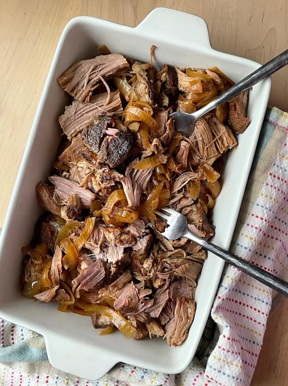 A dish of shredded slow-cooked pulled pork mixed with caramelized onions in a white ceramic baking dish with a serving spoon, placed on a wooden table next to a patterned cloth napkin.