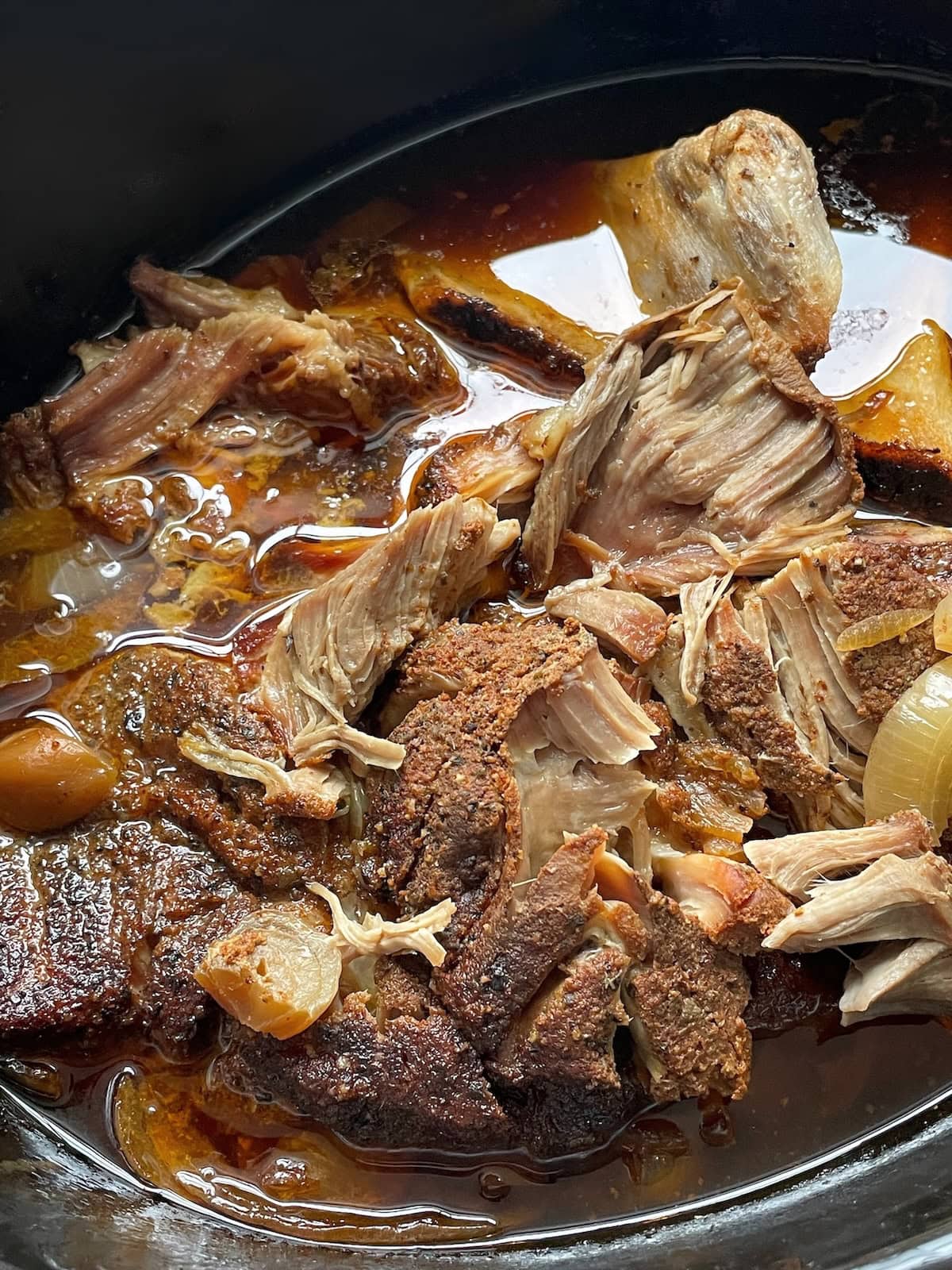 Close-up of shredded pork with spices in a slow cooker, with visible onions and cooking juices.