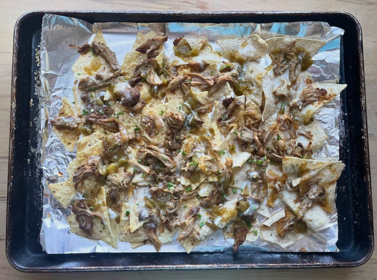 A baking sheet topped with tortilla chips, shredded pork, melted cheese, salsa verde, and diced green chilies.