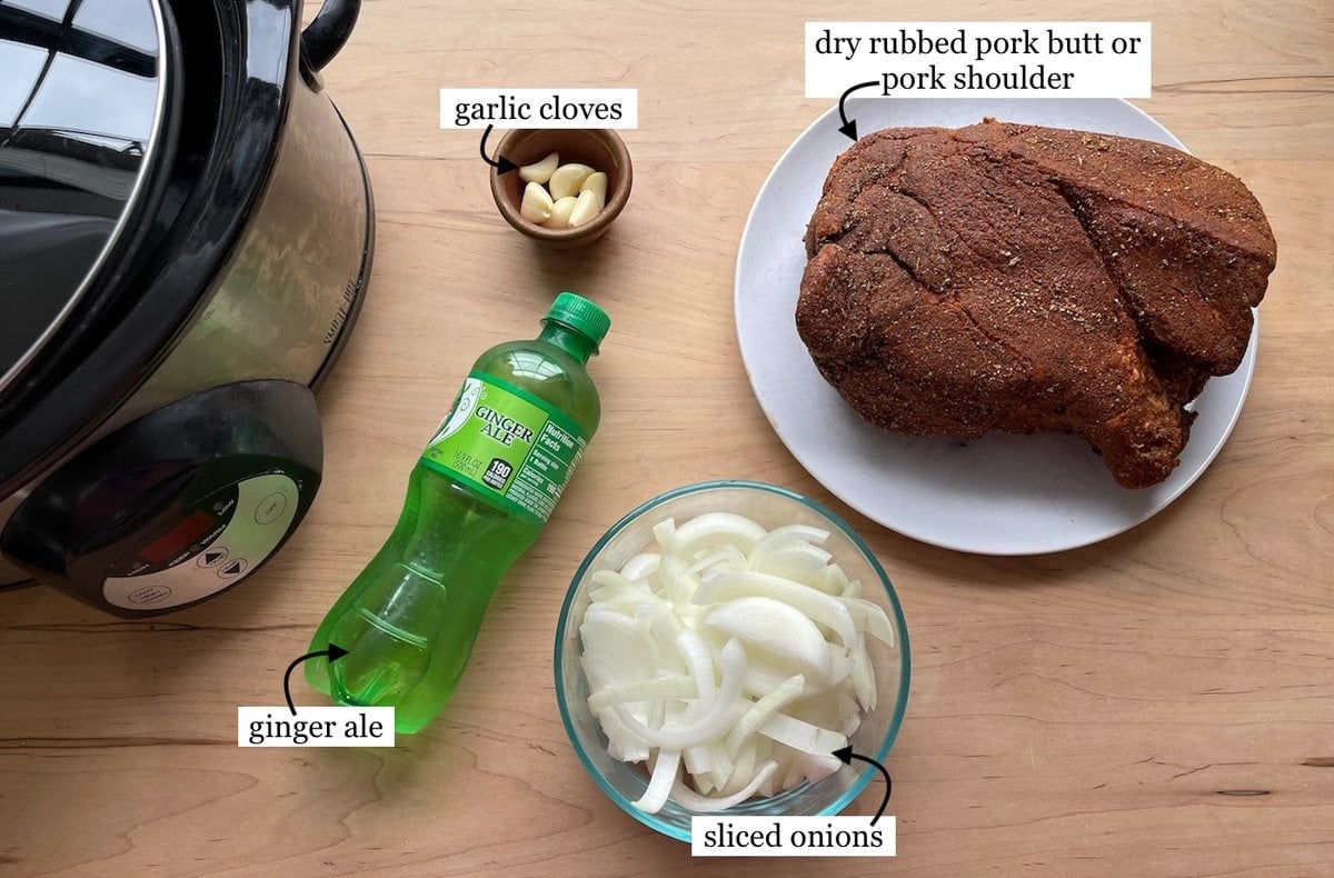 A tabletop view showing ingredients for slow-cooked pork: a dry-rubbed pork butt or pork shoulder, garlic cloves, sliced onions, ginger ale, and a slow cooker.