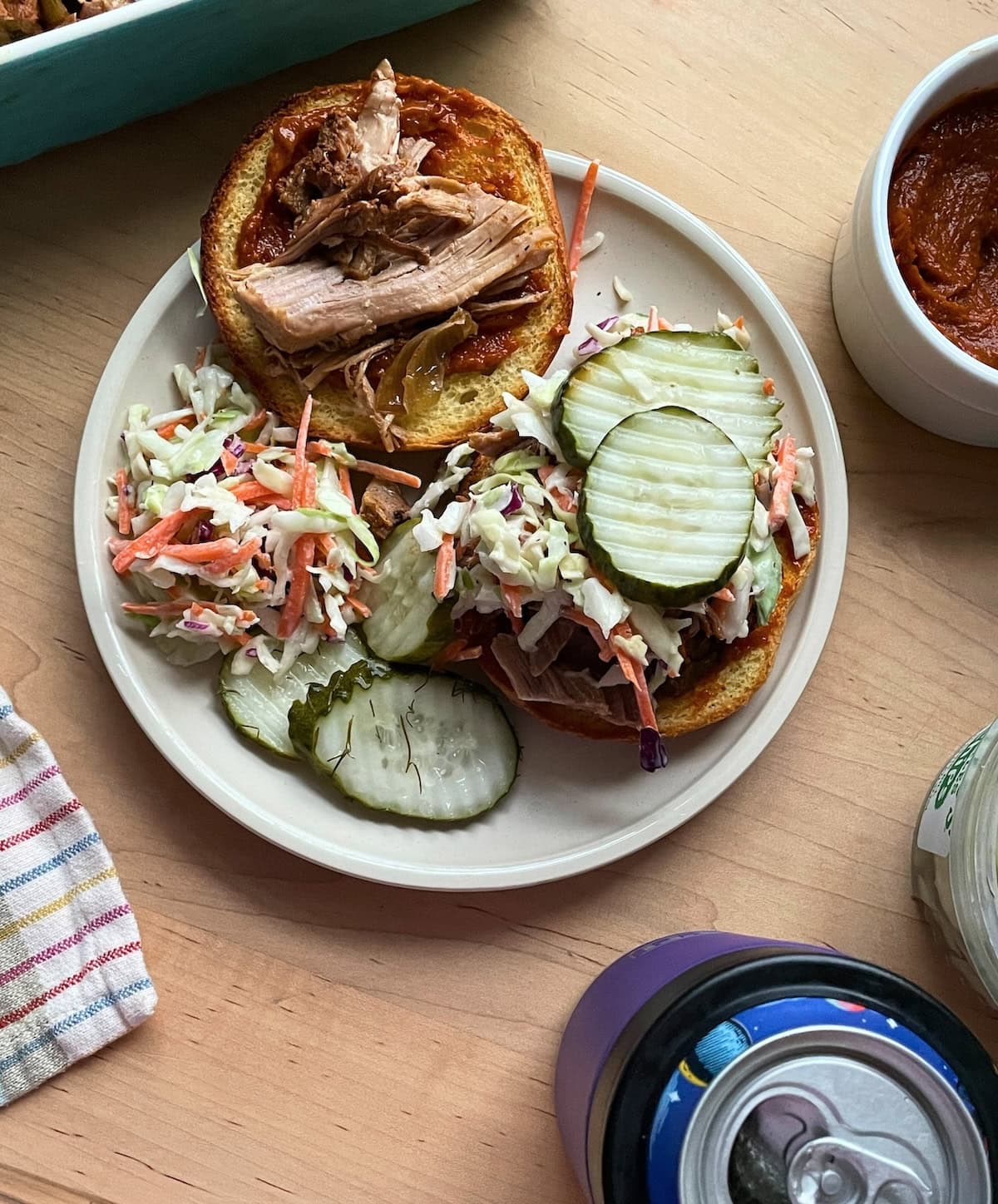 A top view image of an open pulled pork sandwich on a plate with coleslaw and pickles, a bowl of sauce, and a can of beer on a wooden table.