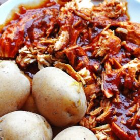 A plate of barbecue pulled chicken with sauce and whole baked potatoes on the side.