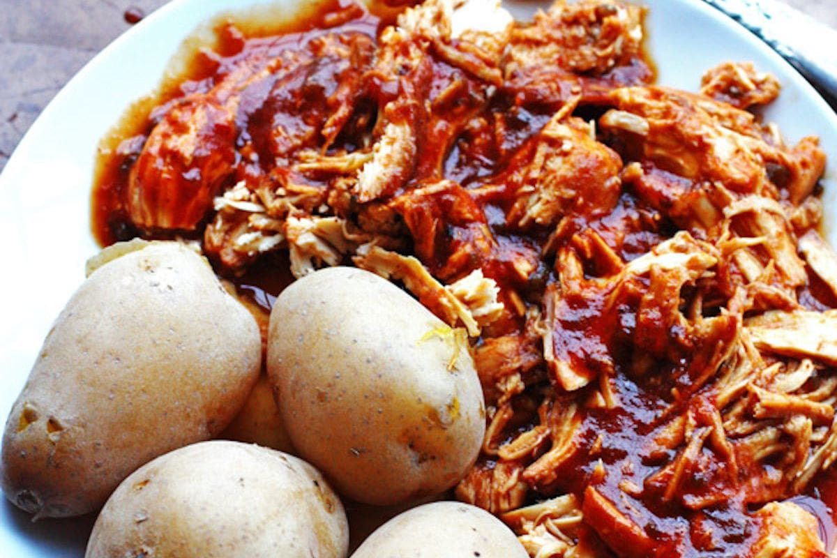 A plate of barbecue pulled chicken with sauce and whole baked potatoes on the side.