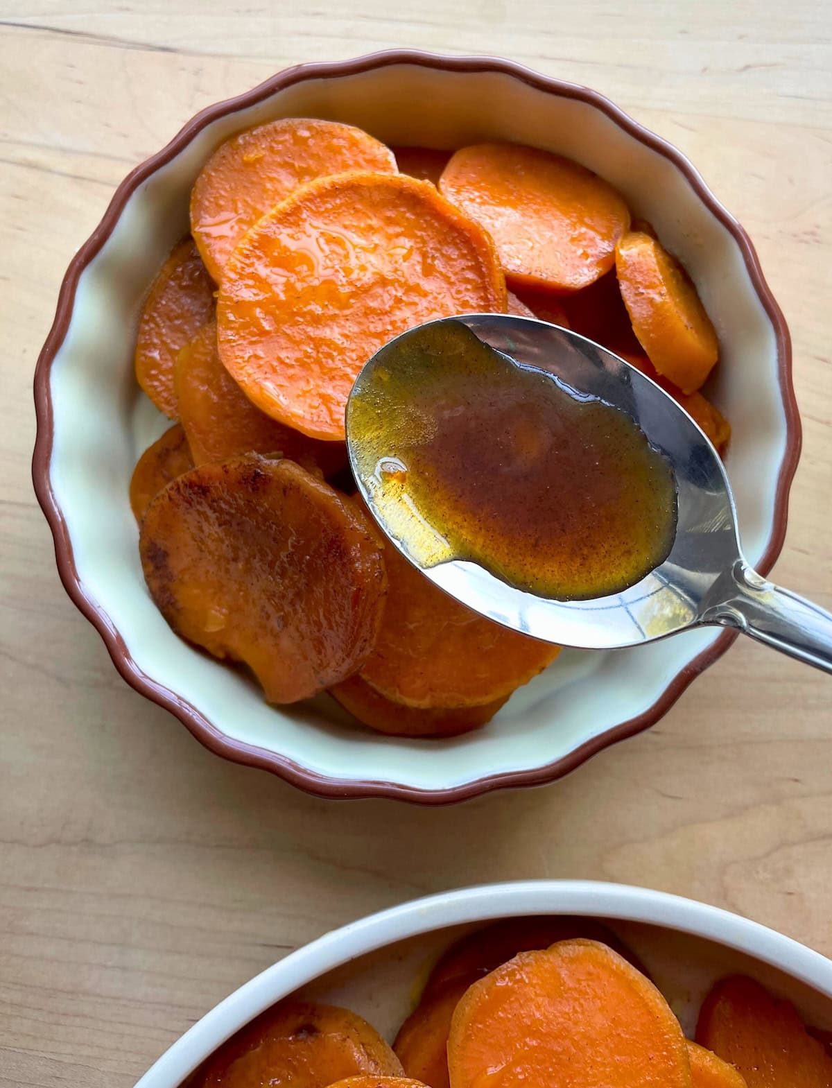 A bowl of sliced sweet potatoes coated in syrup with a spoon, on a wooden surface.