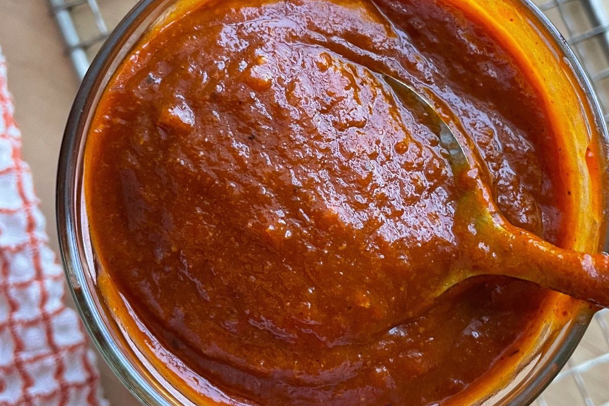 Close-up view of a jar filled with rich, homemade tomato sauce with a spoon inside, set on a kitchen counter beside a checkered cloth.