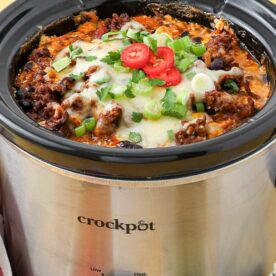 A savory dish of chili topped with melted cheese and green onions in a stainless steel slow cooker.