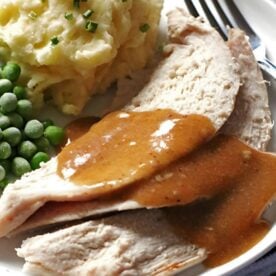 A plate of sliced roast turkey covered in brown gravy, accompanied by mashed potatoes with chives and a serving of green peas.