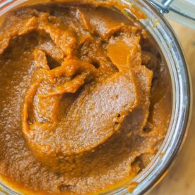 Close-up of pumpkin butter in an open glass jar on a wooden surface, suggesting a homemade ingredient or autumnal recipe component.