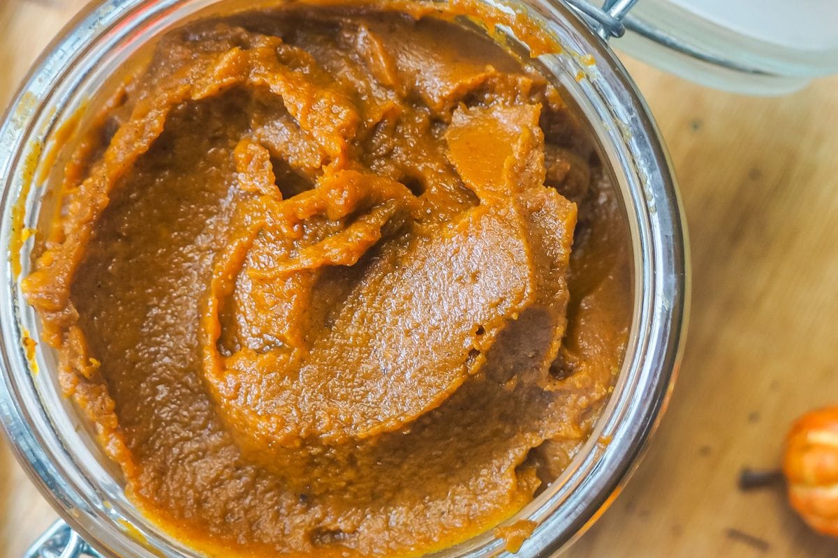 Close-up of pumpkin butter in an open glass jar on a wooden surface, suggesting a homemade ingredient or autumnal recipe component.