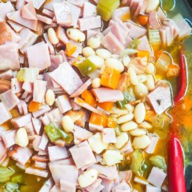 Close-up view of a savory ham and bean soup in a pot with diced ham, beans, onions, celery, and a red chili pepper.