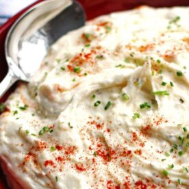 A bowl of homemade mashed potatoes garnished with chopped chives and a sprinkle of paprika, with a serving spoon on the side.
