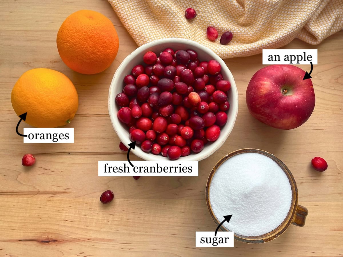 Overhead view of a wooden table with labeled ingredients including oranges, fresh cranberries, an apple, and a bowl of sugar.