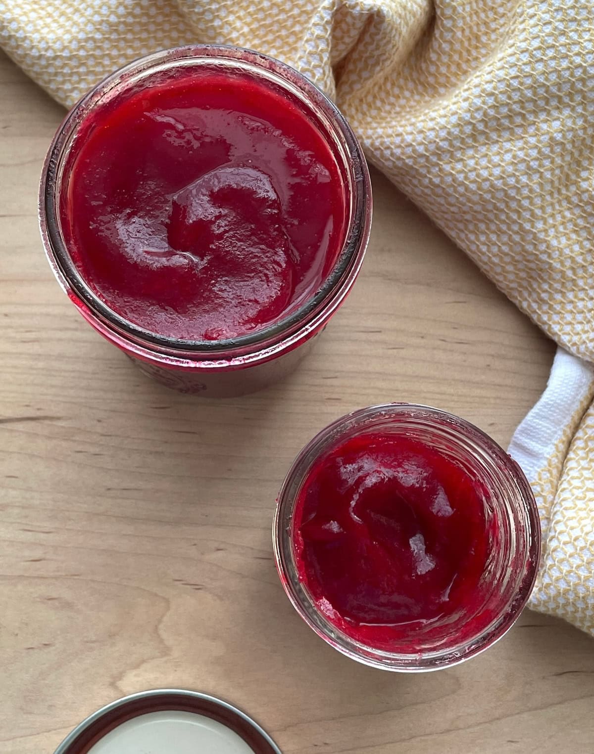 Two open jars filled with bright red cranberry sauce on a wooden surface with a yellow cloth nearby.