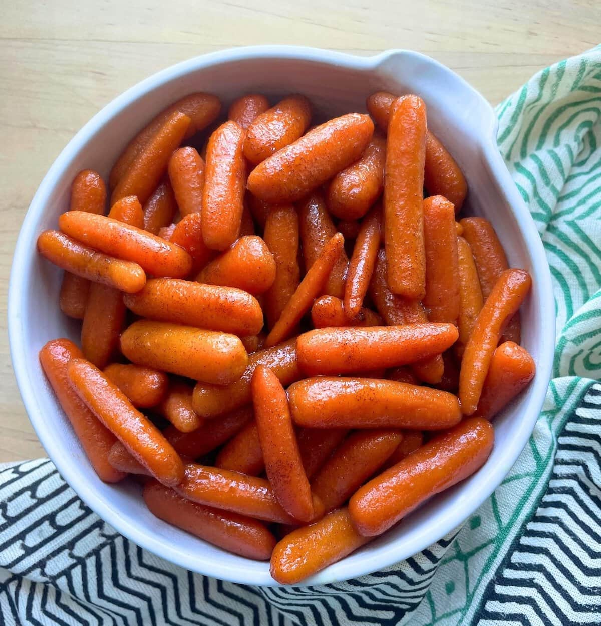 A white bowl full of shiny glazed baby carrots on a wooden surface with a green and white cloth beside it.