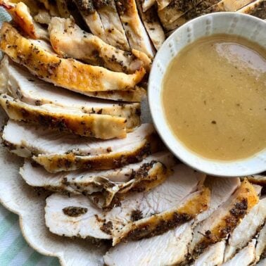 Sliced herb-crusted roast turkey on a plate served with a bowl of gravy.