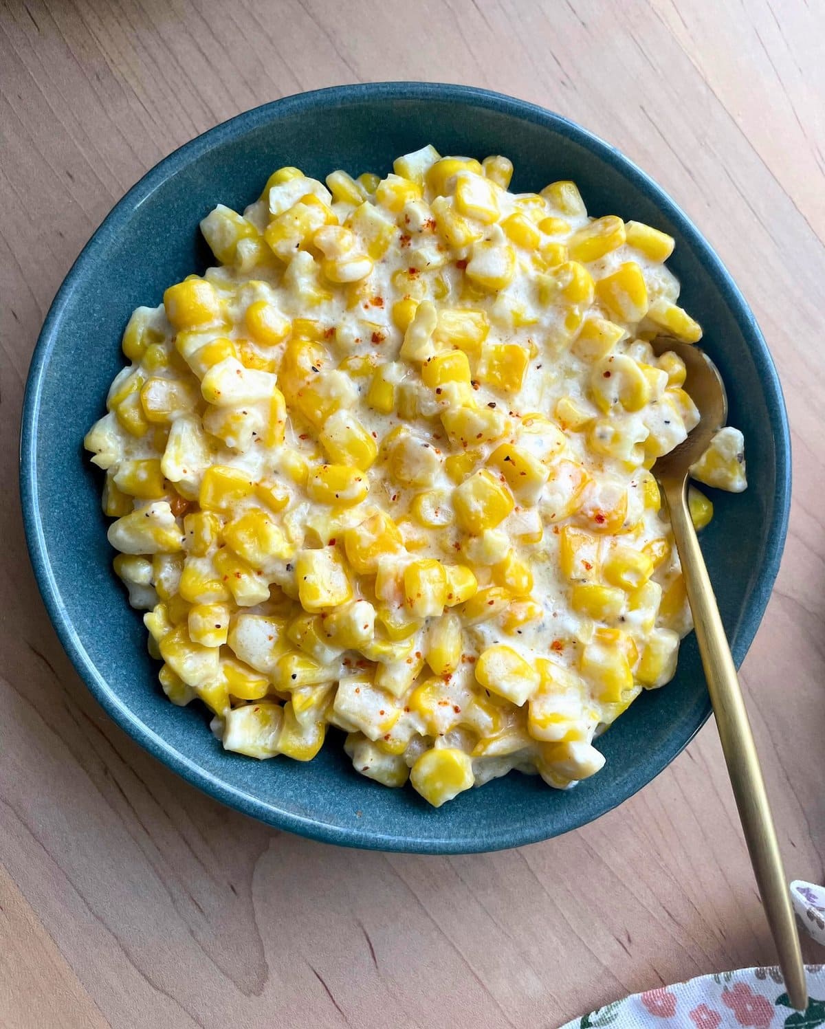 A bowl of creamed corn with a sprinkle of red seasoning on a wooden table, next to a floral cloth napkin.