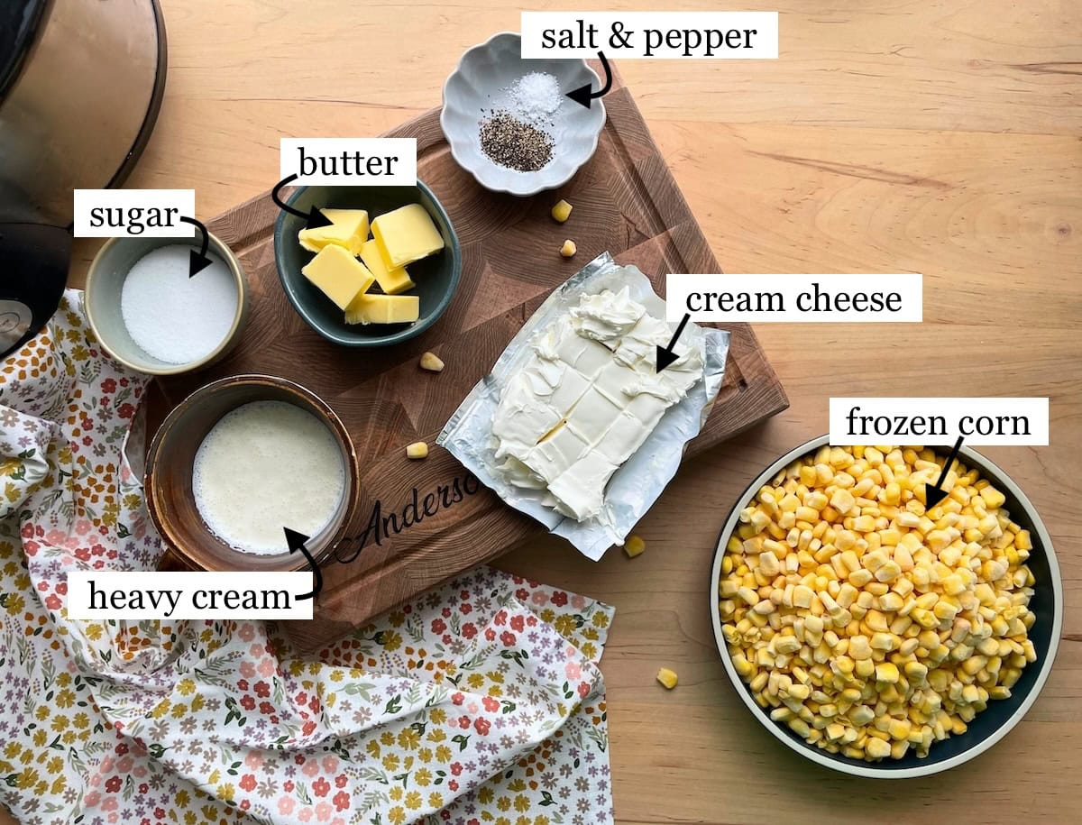 The ingredients in crockpot creamed corn, laid out and labeled on a kitchen counter, including sugar, butter, salt, pepper, cream cheese, heavy cream, and frozen corn.