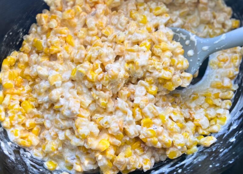 Close-up of a creamy corn casserole with visible corn kernels in a black dish with a gray spatula.