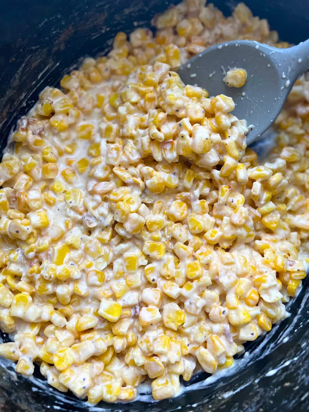 A close-up view of a creamy corn dish in a black crockpot with a gray spatula.