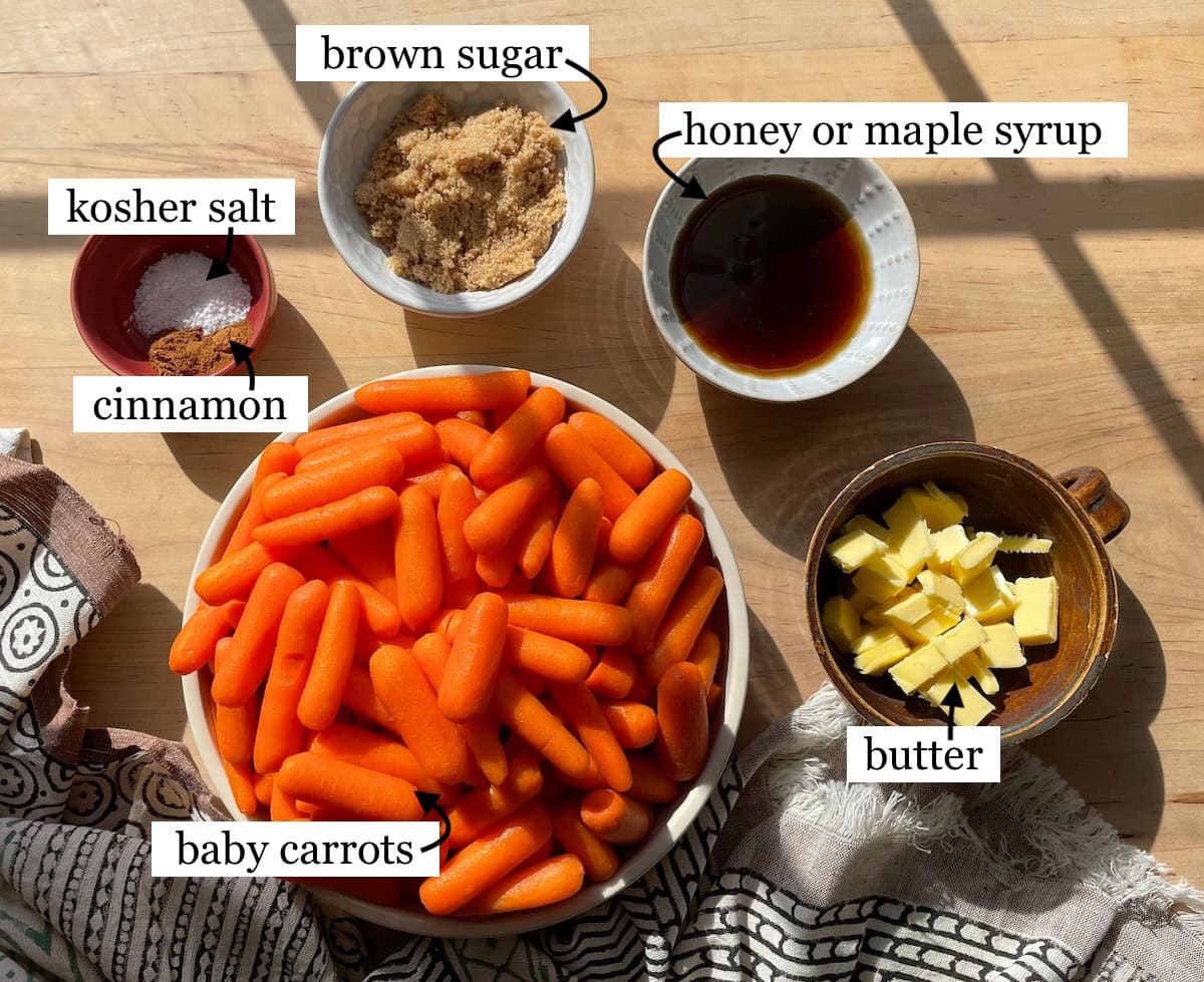 The ingredients for making glazed carrots, including baby carrots, brown sugar, honey or maple syrup, butter, cinnamon, and kosher salt on a wooden countertop with sunlight casting shadows laid out in small bowls and labeled.