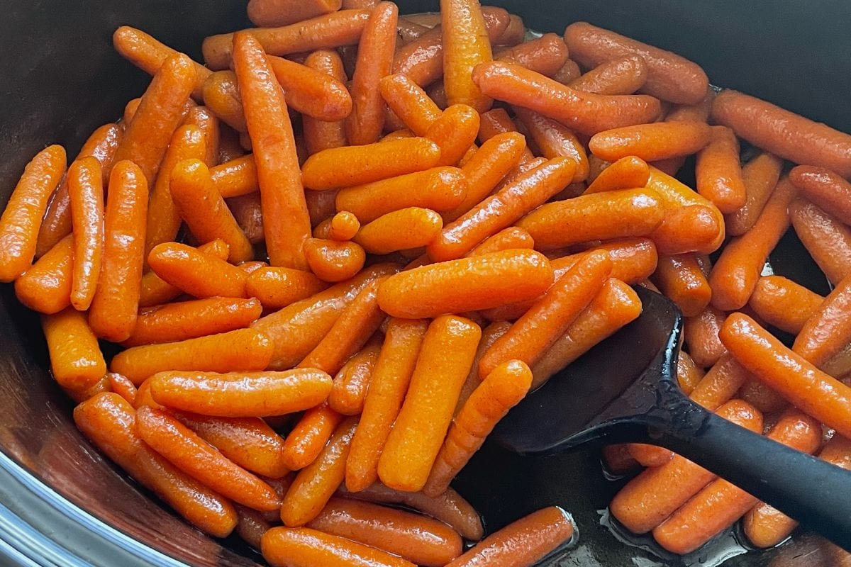 A close-up view of shiny glazed baby carrots in a black cooking pot with a black spatula.