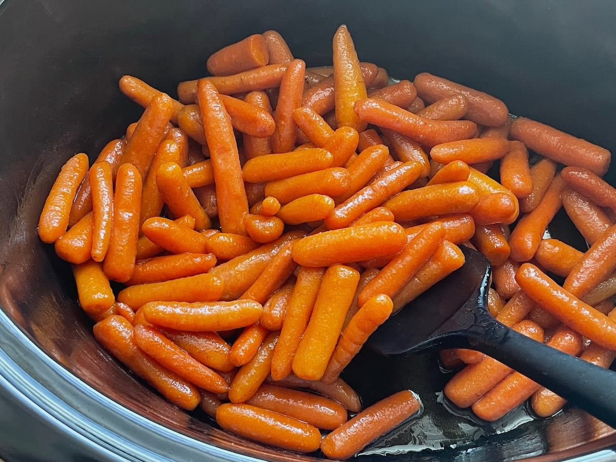 A heap of shiny glazed baby carrots cooked in a slow cooker with a black serving spoon.