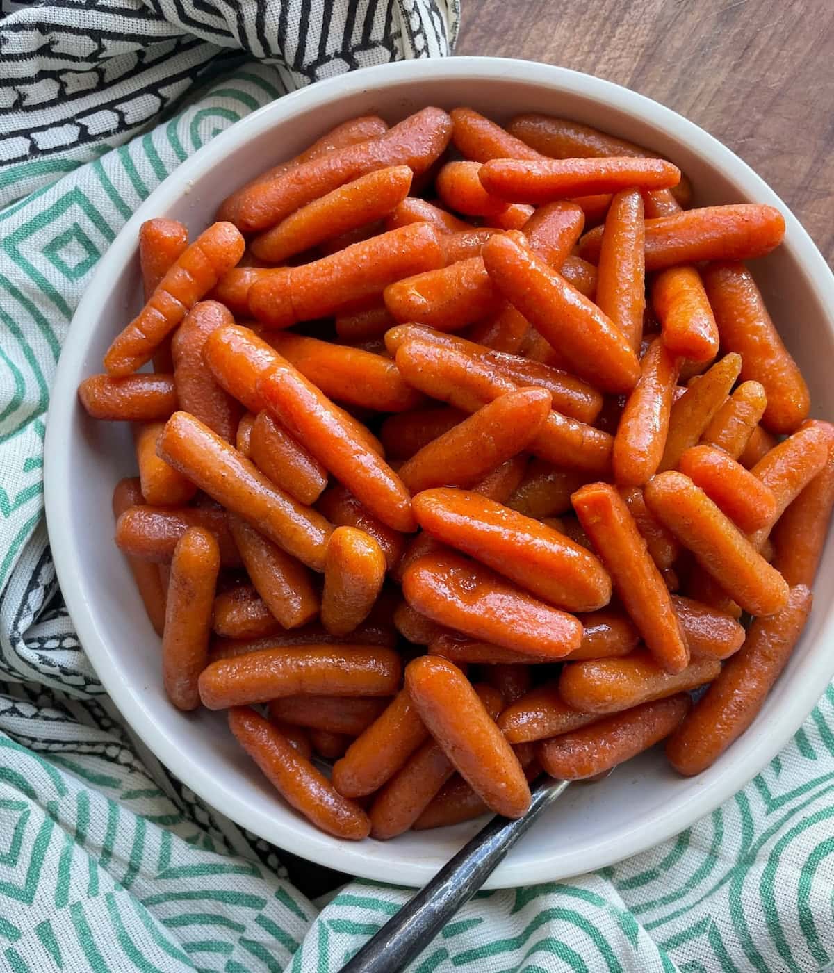 A bowl of shiny glazed baby carrots with a spoon, placed on a patterned cloth, viewed from above.