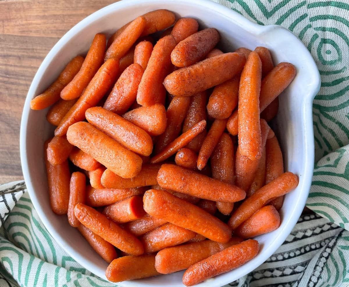 A white bowl filled with orange glazed baby carrots on a green-striped cloth.