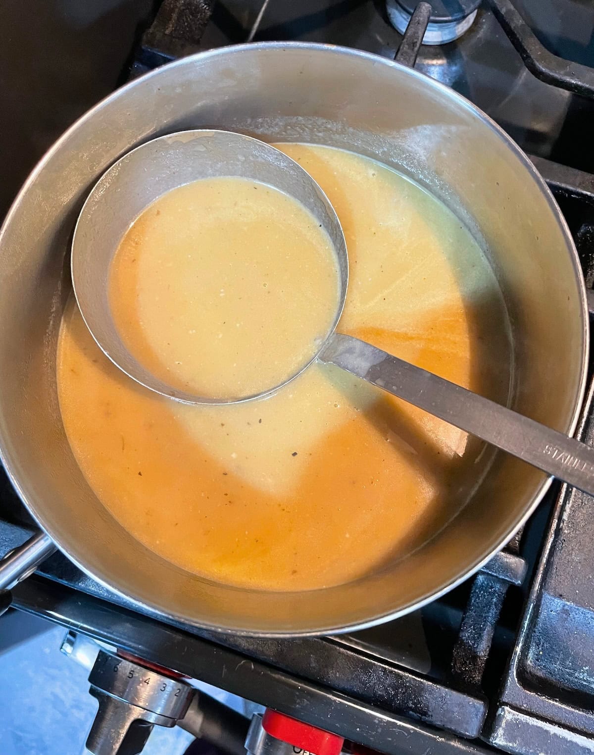 A ladle submerged in a pot of creamy gravy being reduced on a gas stove.