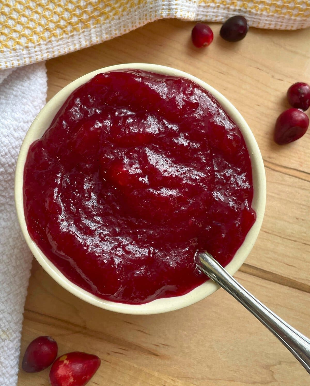 A bowl of cranberry sauce with a spoon on a wooden surface, surrounded by scattered cranberries and a cloth napkin.