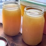Two large glass jars filled with homemade turkey stock, sitting on a wooden surface with a kitchen backdrop.