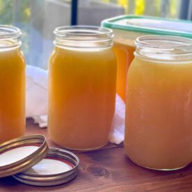 Several jars of homemade broth in varying shades of amber to golden hues, lined up on a wooden surface with lids placed beside them.